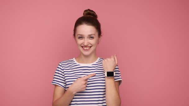 腕時計で腕時計に指を指してパンの髪型を持つ幸せな正の女性は 縞模様のTシャツを着て おもちゃの笑顔でカメラを見て ピンクの背景に隔離された屋内スタジオショット — ストック動画