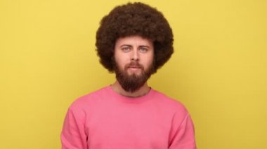 Afro saç stili mutlu sakallı bir adamın portresi parmaklarıyla iyi bir jest yapıyor, işi onaylıyor, kaliteden memnun, pembe kazak giyiyor. Sarı arka planda kapalı stüdyo çekimleri.