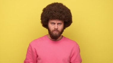 Afro saçlı agresif sakallı adam kameraya kızgın bir ifadeyle bakıyor, aile kavgası, yanlış anlaşılma, pembe kazak giyiyor. Sarı arka planda kapalı stüdyo çekimleri.