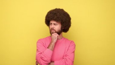 Afro saçlı dalgın bir adam çenesini elleriyle tutuyor, düşünme ihtiyacı duyuyor, strateji planlıyor, başlangıç için fikirler düşünüyor, pembe kazak giyiyor. Sarı arka planda kapalı stüdyo çekimleri.
