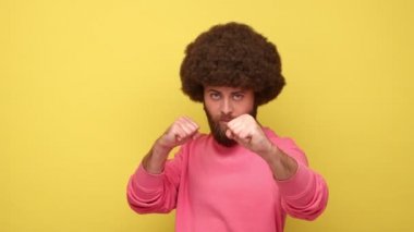 Afro saçlı sakallı adam boks jesti ile ayakta duruyor, kameraya yumruk atıyor, kavgayı kışkırtıyor ya da kendini savunmaya hazır pembe kazak giyiyor. Sarı arka planda kapalı stüdyo çekimleri.