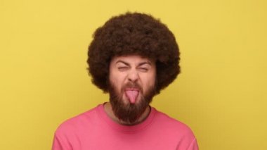 Afro saçlı, çocuksu sakallı, yaramaz, itaatsiz, suratını buruşturan, pembe kazak giyen bir adam. Sarı arka planda kapalı stüdyo çekimleri.