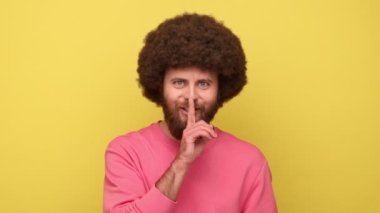 Afro saç stili iyimser bir adam sessiz bir jest yapıyor, parmaklarını dudaklarının yanında tutuyor ve gülümsüyor, sırlarını saklamaya devam ediyor, pembe kazak giyiyor. Sarı arka planda kapalı stüdyo çekimleri.