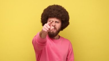Afro saçlı sakallı adam kameraya kızgın bir ifadeyle bakıyor, seni suçluyor, seni işaret ediyor, seçim yapıyor, pembe kazak giyiyor. Sarı arka planda kapalı stüdyo çekimleri.