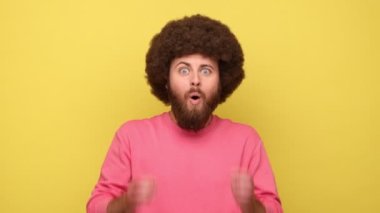 Afro saçlı heyecanlı sakallı adam hayret verici bir ifadeyle kameraya bakıyor, hoş bir sürpriz, pembe kazak giyiyor. Sarı arka planda kapalı stüdyo çekimleri.