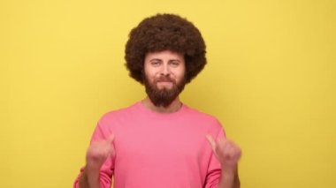 Sakallı, Afro saçlı, kendini gösteren ve kameraya bakan dişlek gülüşlü, son derece memnun pembe kazak giyen bir adam. Sarı arka planda kapalı stüdyo çekimleri.