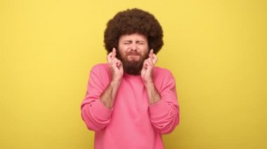 Afro saç stili parmakları kesişen sakallı adam, gözlerini kapayan yalvaran yüzüyle dilek tutan pembe kazak giyen neşeli rüya gerçekleşiyor. Sarı arka planda kapalı stüdyo çekimleri.