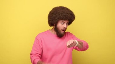Sakallı, Afro saçlı bir adam kol saatine bakıyor, tırnaklarını yiyor, saati kontrol ediyor, geç kalıyor, son teslim tarihi, pembe kazak giyiyor. Sarı arka planda kapalı stüdyo çekimleri.