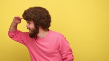 Afro saç stili sakallı bir adam ellerini gözlerine uzatıp pembe kazak giymiş, el sallayarak ufuk çizgisini seyrediyor. Sarı arka planda kapalı stüdyo çekimleri