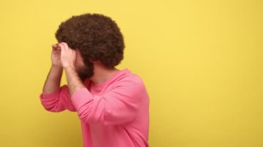 Sakallı, Afro saçlı, gözlerinin yanında yumruklar tutan dürbün hayal eden, deliklerden bakan, gözetleyen, eğlenen, pembe kazak giyen bir adam. Sarı arka planda kapalı stüdyo çekimleri.