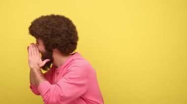 Sakallı, sinirli, Afro saç stili yüksek sesle bağıran, ağzı yakınında el ele tutuşan, protesto eden, pembe kazak giyen sinirli bir adam. Sarı arka planda kapalı stüdyo çekimleri.