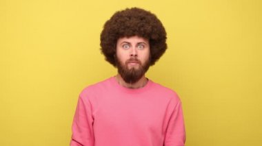 Sakallı adam, Afro saç stili, ağzı ağızla kaplı, korkmuş şok olmuş gözlerle bakıyor, gizli tabuyu koruyor, pembe kazak giyiyor. Sarı arka planda kapalı stüdyo çekimleri.