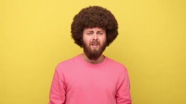 Afro saç stili sakallı bir adam yüzünde yüz makyajı ile duruyor, kendini suçluyor, kötü anılar yüzünden pişmanlık duyuyor, pembe kazak giyiyor. Sarı arka planda kapalı stüdyo çekimleri.