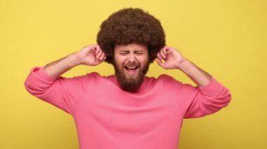 Sakallı, rahatsız edici, Afro saçlı, parmaklarıyla kulaklarını kapatan, rahatsız edici seslerden bıkmış, sessizlik isteyen, pembe kazak giyen bir adam. Sarı arka planda kapalı stüdyo çekimleri.