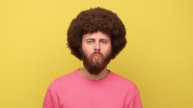 Afro saçlı, dudak büken balık suratlı, komik çocuksu suratlı, dişlek gülüşlü, pembe kazaklı kameraya bakan bir adam. Sarı arka planda kapalı stüdyo çekimleri.
