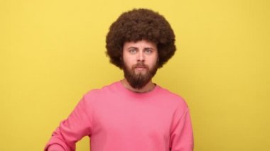 Afro saçlı sakallı adam kameraya bakıyor, parmak tabancasını kafasına doğrultuyor, tabancayla kendini vuruyor, pembe kazak giyiyor. Sarı arka planda kapalı stüdyo çekimleri.