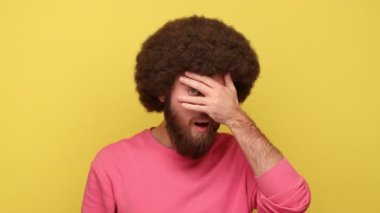 Sakallı, Afro saçlı, parmakları delik deşik eden, gözleri kollarıyla kapayan, gülümseyen, gözetleyen pembe kazak giyen bir adam. Sarı arka planda kapalı stüdyo çekimleri.