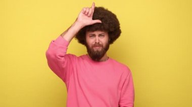 Afro saçlı sakallı bir adam alnına yakın bir yerde parmaklarını tutarken gösteriyor. Üzücü bir hata, pembe kazak giyiyor. Sarı arka planda kapalı stüdyo çekimleri.