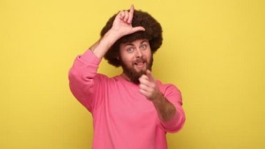 Sen daha eziksin. Afro saçlı sakallı adam ezik jestleri yapıyor ve kamerayı işaret ediyor, yenilgiyle dalga geçiyor, pembe kazak giyiyor. Sarı arka planda kapalı stüdyo çekimleri.