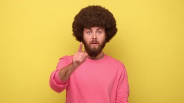 Afro saçlı sakallı adam kameraya bakıyor ve başka bir şekilde işaret ediyor, patronvari bir ifadeyle ayrılmasını emrediyor, pembe kazak giyiyor. Sarı arka planda kapalı stüdyo çekimleri.