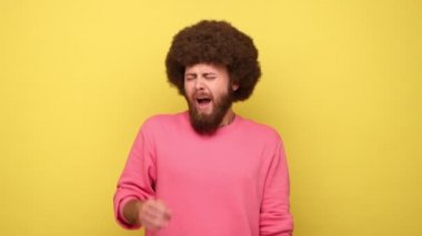 Sakallı, uykusuz, Afro saç stili esneyen, ellerini kaldıran, fazla mesai yapmaktan yorgun hisseden pembe kazak giyen bir adam. Sarı arka planda kapalı stüdyo çekimleri.