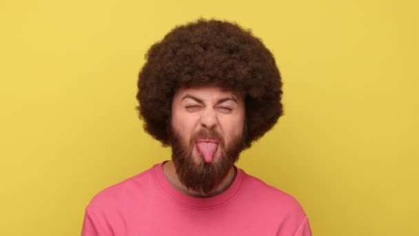 アフリカの髪型をした子供の髭の男は ピンクのスウェットシャツを着て 顔を作って いたずらな不従順な優雅さで舌を見せました 黄色の背景に隔離された屋内スタジオショット — ストック動画