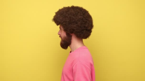 留着非洲式胡子的男人转过头对着相机 愉快地挥挥手 打招呼 表达幸福 穿着粉色运动衫 在黄色背景下隔离的室内拍摄 — 图库视频影像