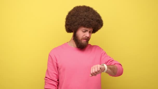 髭を生やした思慮深い男とアフロの髪型は ピンクのトレーナーを着て 期限を心配し 彼のスマートウォッチを見て混乱した 黄色の背景に隔離された屋内スタジオショット — ストック動画
