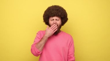 Afro saçlı sakallı bir adam avuç içlerine uzanmış ve mutlu bir şekilde gülümsüyor. Rahat bir şekilde uyuyor, dinleniyor, uyukluyor ve pembe bir kazak giyiyor. Sarı arka planda kapalı stüdyo çekimleri.