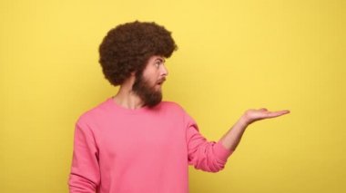Afro saç stili sakallı, afro sakallı, fotokopi çekilen, ticari metin ve eşyalar için boş bir yer gösteren, pembe kazaklı bir adam. Sarı arka planda kapalı stüdyo çekimleri.