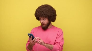 Afro saçlı sakallı hippi adam cep telefonunu boş ekran ile işaret ediyor. Yüzünde şaşırmış bir ifade var. Pembe kazak giyiyor. Sarı arka planda kapalı stüdyo çekimleri.