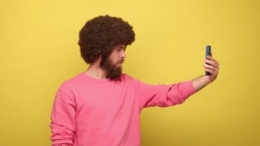 Sakallı hippi pozu veren, Afro saç stili pozu veren, akıllı telefondan özçekim yapan, alet kamerasına bakan, blog yazan, pembe kazak giyen bir adam. Sarı arka planda kapalı stüdyo çekimleri.