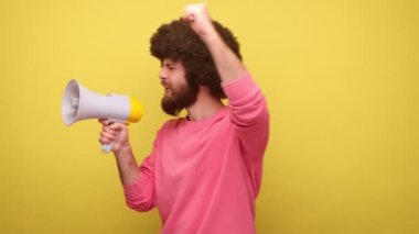 Afro saç stili protestolu sakallı adam elinde megafonla bağırarak konuşuyor, önemli bir mesaj veriyor, pembe kazak giyiyor. Sarı arka planda kapalı stüdyo çekimleri.