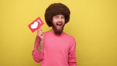 Afro saçlı, mutlu gülüşlü sakallı bir adam, sosyal medyayı işaret eden ikon gibi, kalp düğmesine basan, en iyi blog içeriğini seçen, kazak giyen. Sarı arka planda kapalı stüdyo çekimleri.