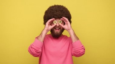 Sakallı hippi sakallı, Afro saç stili, altın bitcoin ile kaplı, engelleme zinciri konsepti, olumlu duygularını ifade eden, pembe kazak giyen. Sarı arka planda kapalı stüdyo çekimleri.
