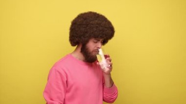 Sakallı hippi sakallı, Afro saçlı, camdan soğuk su içen, dişlerinde güçlü bir ağrı hisseden, yanağına dokunan, pembe kazak giyen bir adam. Sarı arka planda kapalı stüdyo çekimleri.