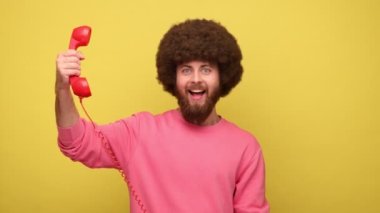 Sakallı hippi adam, Afro saç stili tutuyor ve kameraya cep telefonunu gösteriyor, eski telefon aracılığıyla hoş sohbetler ediyor, pembe kazak giyiyor. Sarı arka planda kapalı stüdyo çekimleri.