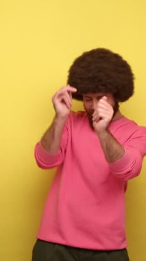 Afro saç stili sakallı bir adamın hızlı ritimli, başarıyı kutlayan, pembe kazak giyen favori şarkısını beğendiği dikey video görüntüleri. Sarı arka planda kapalı stüdyo çekimleri.