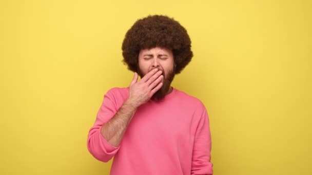 アフロのヘアスタイルを持つひげを生やした男が手のひらの上に寝そべって笑って満足し 快適な昼寝をして うとうとし ピンクのスウェットシャツを着ている 黄色の背景に隔離された屋内スタジオショット — ストック動画