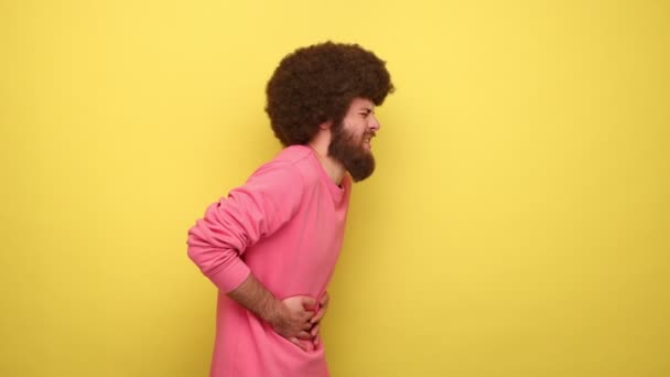 アフリカの髪型を持つ不健康な男の側面図は 胃の痛みを感じます 虫垂炎の下痢や便秘のリスクを受けます ピンクのトレーナーを身に着けています 黄色の背景に隔離された屋内スタジオショット — ストック動画