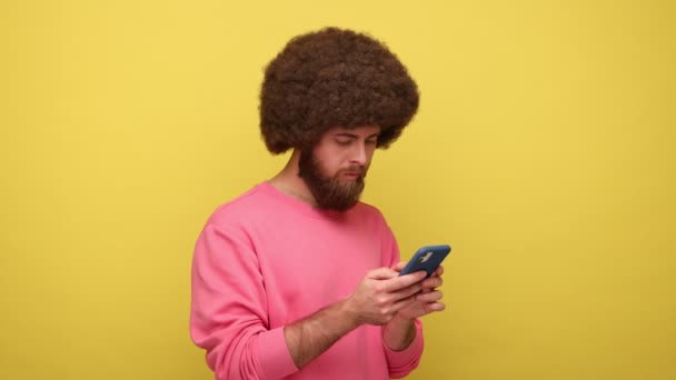 带着快乐笑容的嬉皮士男人 有着非洲式的发型 用手机进行在线交流 打字按摩 穿粉色运动衫 在黄色背景下隔离的室内拍摄 — 图库视频影像