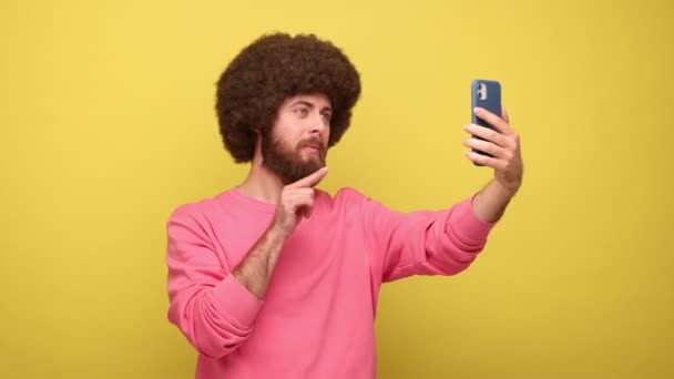 アフロヘアスタイルの放送のライブストリームを持つ髭ヒップスター男性は フォロワー 広告と話をし 購読するように求めて ピンクのスウェットシャツを着ている 黄色の背景に隔離された屋内スタジオショット — ストック動画