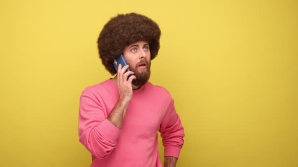 留着胡子 留着非洲式发型的嬉皮士男人打电话给亲戚或朋友聊天 保持友好的对话和微笑 穿着粉色运动衫 在黄色背景下隔离的室内拍摄 — 图库视频影像