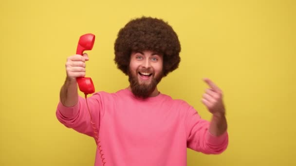 戴着非洲发式手指的男人 手握红色复古电话 等你的电话 看着相机 穿着粉色运动衫 在黄色背景下隔离的室内拍摄 — 图库视频影像