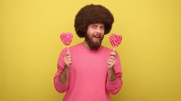 アフロの髪型の髭ヒップスター空腹の男は ピンクのスウェットシャツを着て お菓子を味わいたい キャンディをなめる 心の形で2つのLolypopsを保持している 黄色の背景に隔離された屋内スタジオショット — ストック動画