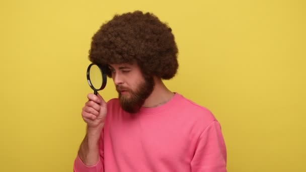 虫眼鏡を保持し 大きなズームの目でカメラを見て ピンクのスウェットシャツを着て アフロの髪型を持つ髭ヒップスター男性 黄色の背景に隔離された屋内スタジオショット — ストック動画