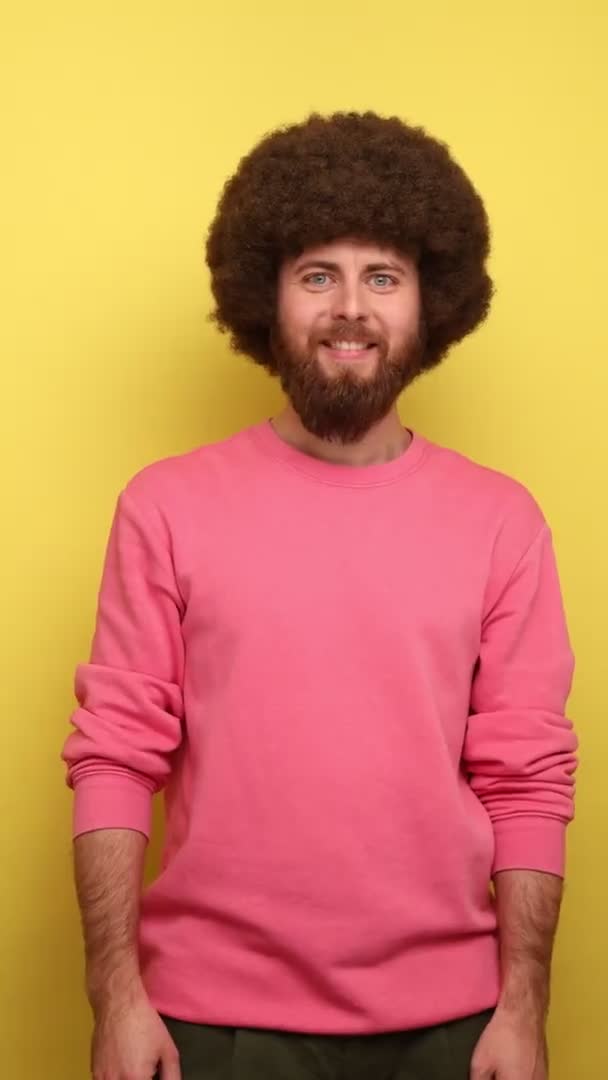 一个有着非洲式发型的男人站在镜头前 面带微笑地看着相机 朝下看 主动提出要刷牙 穿着粉色运动衫 在黄色背景下隔离的室内拍摄 — 图库视频影像