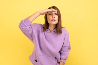 Dikkatle hazırlanmış güzel bir kadının portresi. Ciddi bir şekilde ayakta duruyor, eli alnında ve uzaklara bakıyor, mor kapüşonlu bir şey giyiyor. Sarı arka planda kapalı stüdyo çekimleri.