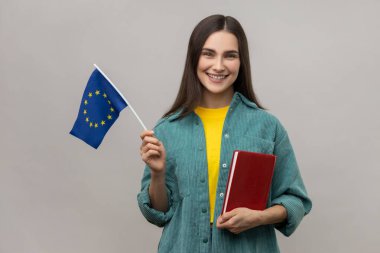 Elinde kitap ve Avrupa Birliği bayrağı olan, yurtdışında eğitim gören, kameraya gülümseyen, günlük tarz ceket giyen pozitif öğrenci kadın portresi. Gri arka planda izole edilmiş kapalı stüdyo görüntüleri.