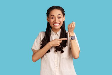 Siyah rastalı mutlu kadının portresi, kameraya bakıyor, akıllı saatini gösteriyor, beyaz gömlek giyiyor. Mavi arka planda kapalı stüdyo çekimi.